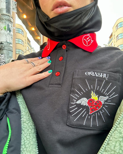 A person wearing a black jacket with a red collar and buttons. The jacket has a patch on the left chest that reads ‘OMASHU’ with a heart and a bird. The person is also wearing a black scarf around their neck and multiple rings on their fingers. The background is a city street with buildings and a lamppost.