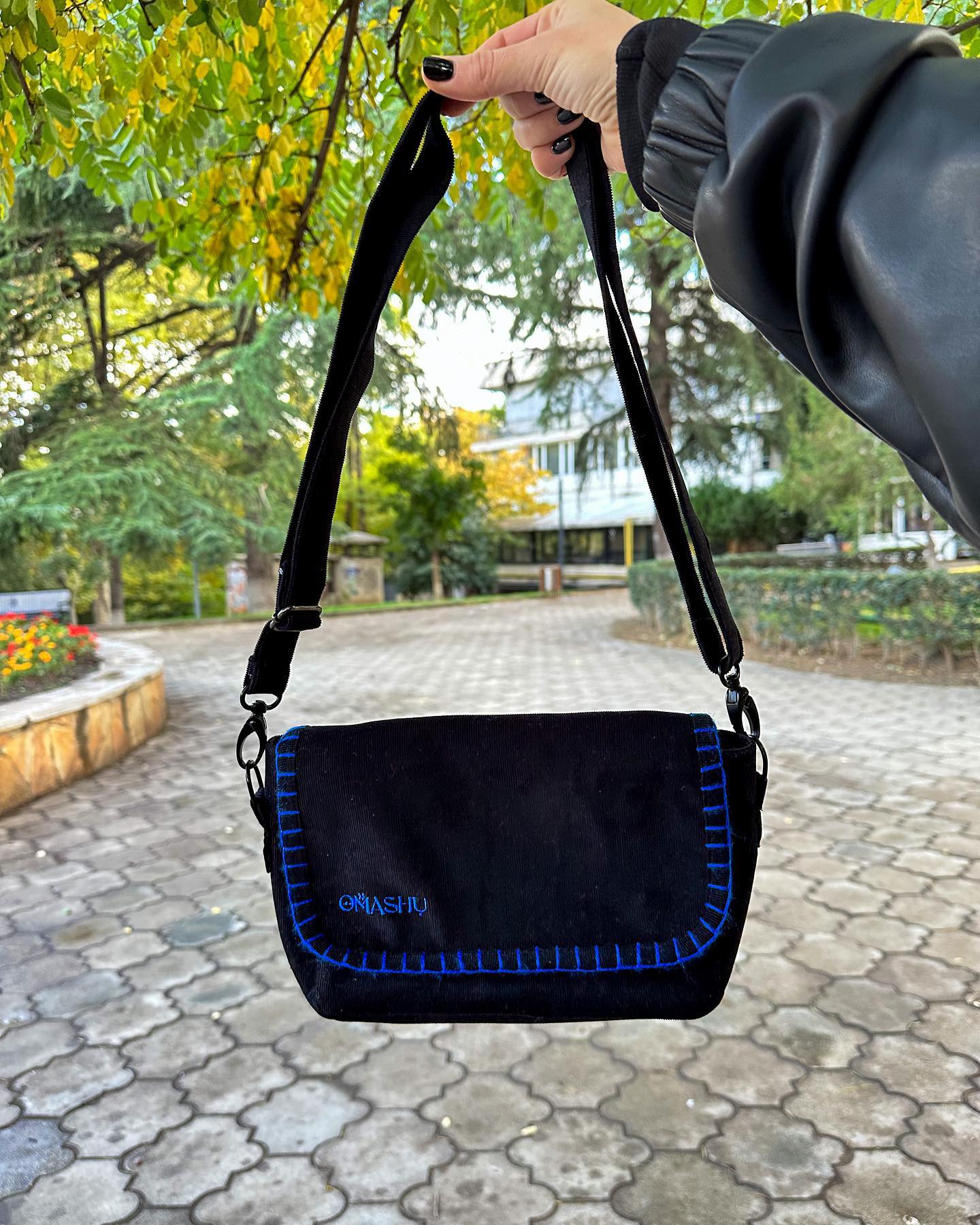 A photo of a black OMASHU crossbody bag with blue stitching, being held by a hand with black nail polish, featuring a black strap with silver hardware and a blue logo on the front that reads ‘OMASHU’, against a cobblestone street background with trees and a building, taken during the day.