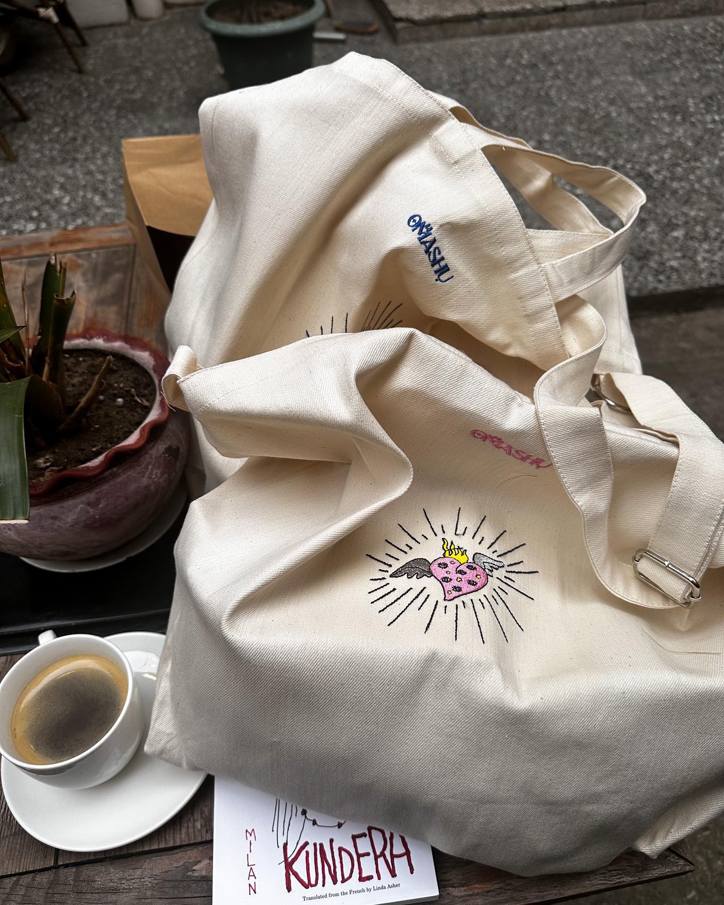 A white Omashu tote bag with a pink and black graphic of a heart with rays coming out of it, resting on a wooden table with a potted plant and a cup of coffee next to it, and a white paper with ‘KUNDERA’ written on it in black ink, against a concrete floor background with a wooden bench.