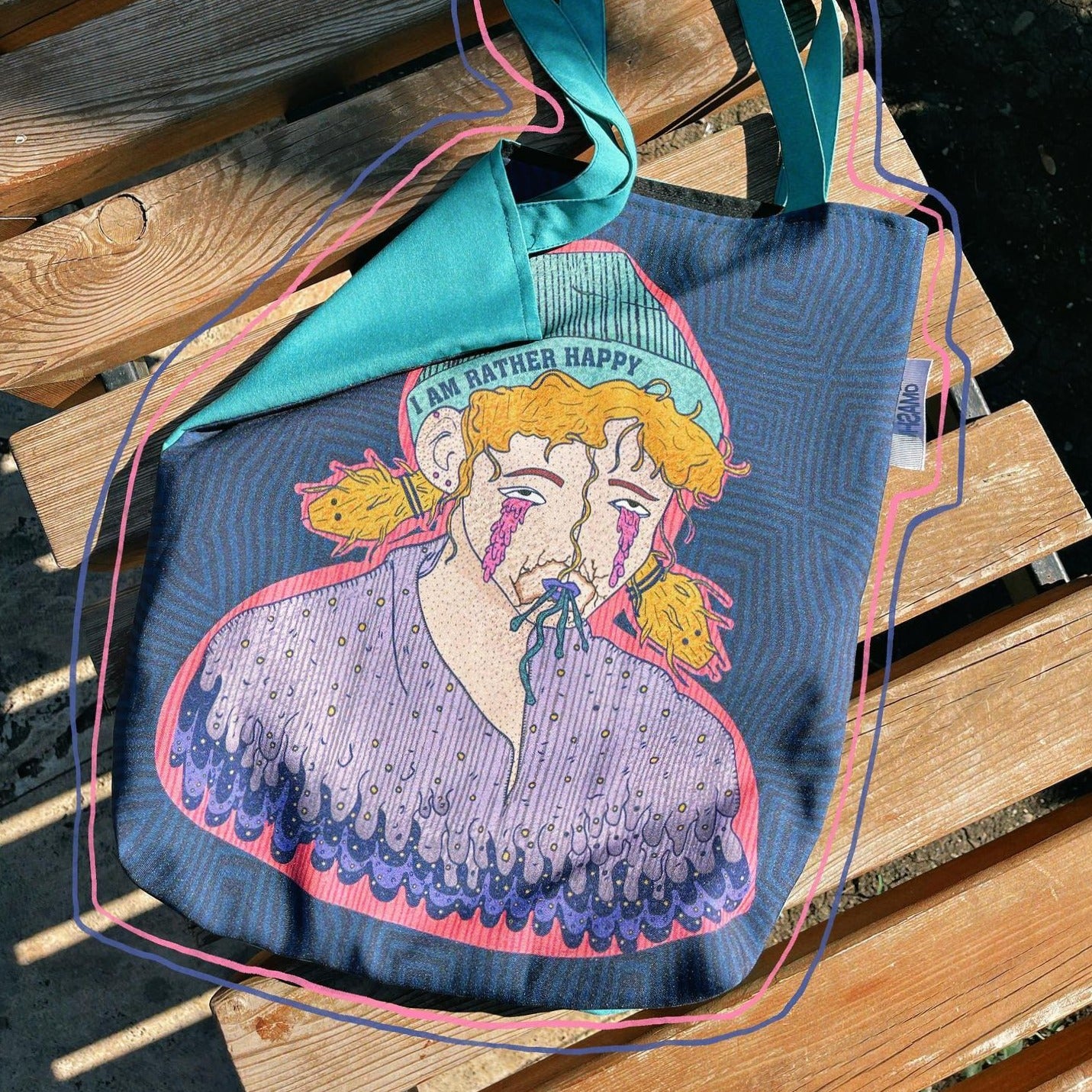 A blue Omashu tote bag with a colorful design of a person’s face with pink hair and a blue nose, wearing a purple shirt with a pink collar, with a smile on their face, and text above their head that reads ‘I am rather happy’, with two teal straps, resting on a wooden bench on a sunny day with a tree and a building in the background.