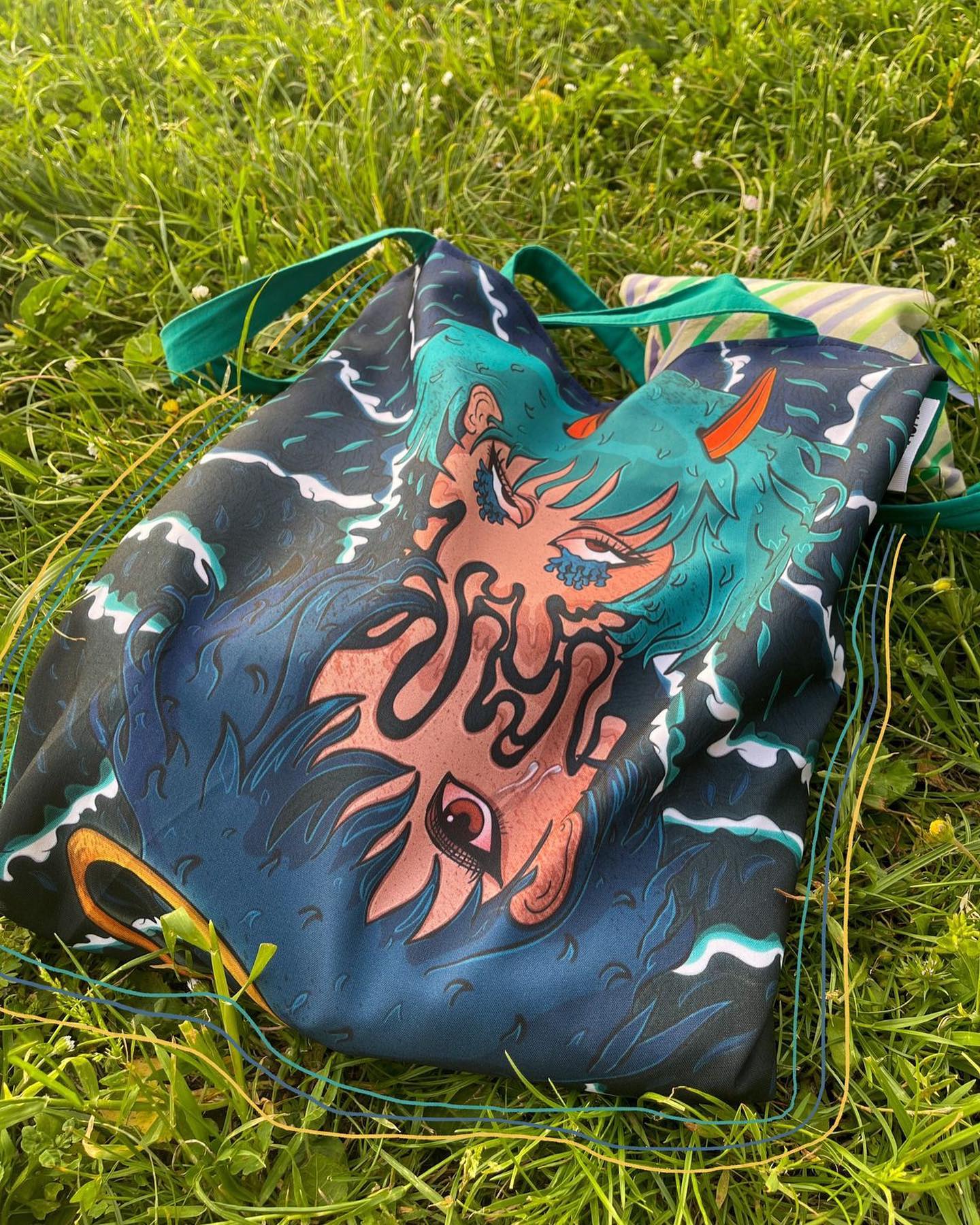 A tote bag lying on a grassy field. The tote bag is made of a shiny material and has a colorful design on it. The design is an abstract illustration of a dragon in shades of blue, green, and orange. The bag has two green straps and a small orange tag on the side. The grass is bright green and appears to be freshly cut.