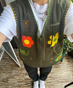 Person wearing green vest with yellow trim and green zipper down middle with red flower, yellow flower, and cat face patches on it with white shirt, black pants, and white sneakers standing on wooden deck with potted plant .