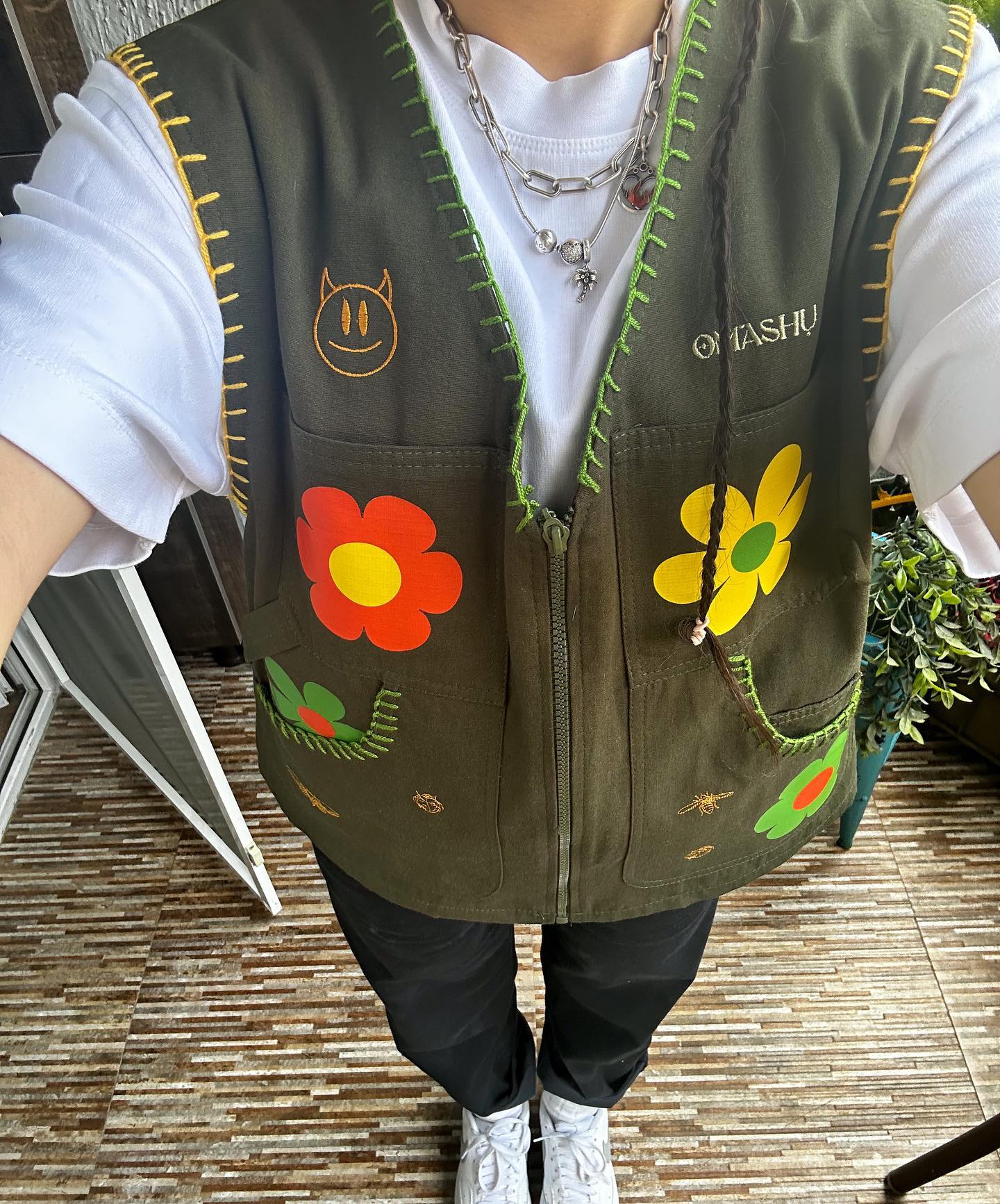 Person wearing green vest with yellow trim and green zipper down middle with red flower, yellow flower, and cat face patches on it with white shirt, black pants, and white sneakers standing on wooden deck with potted plant .