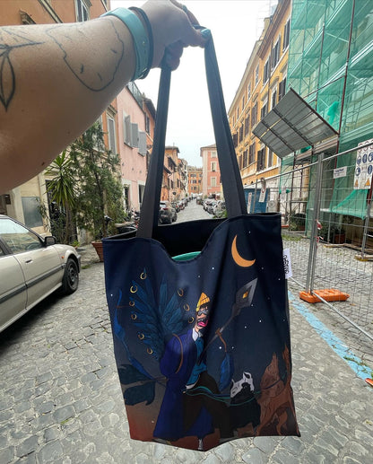 A photo of a person holding a black Omashu tote bag with a colorful design featuring a night sky with a crescent moon, a tree with blue leaves, and a cat sitting on a branch, on a cobblestone street in a European city, possibly Rome, Italy, with buildings and cars parked on the street in the background and a tattoo on the person’s arm.