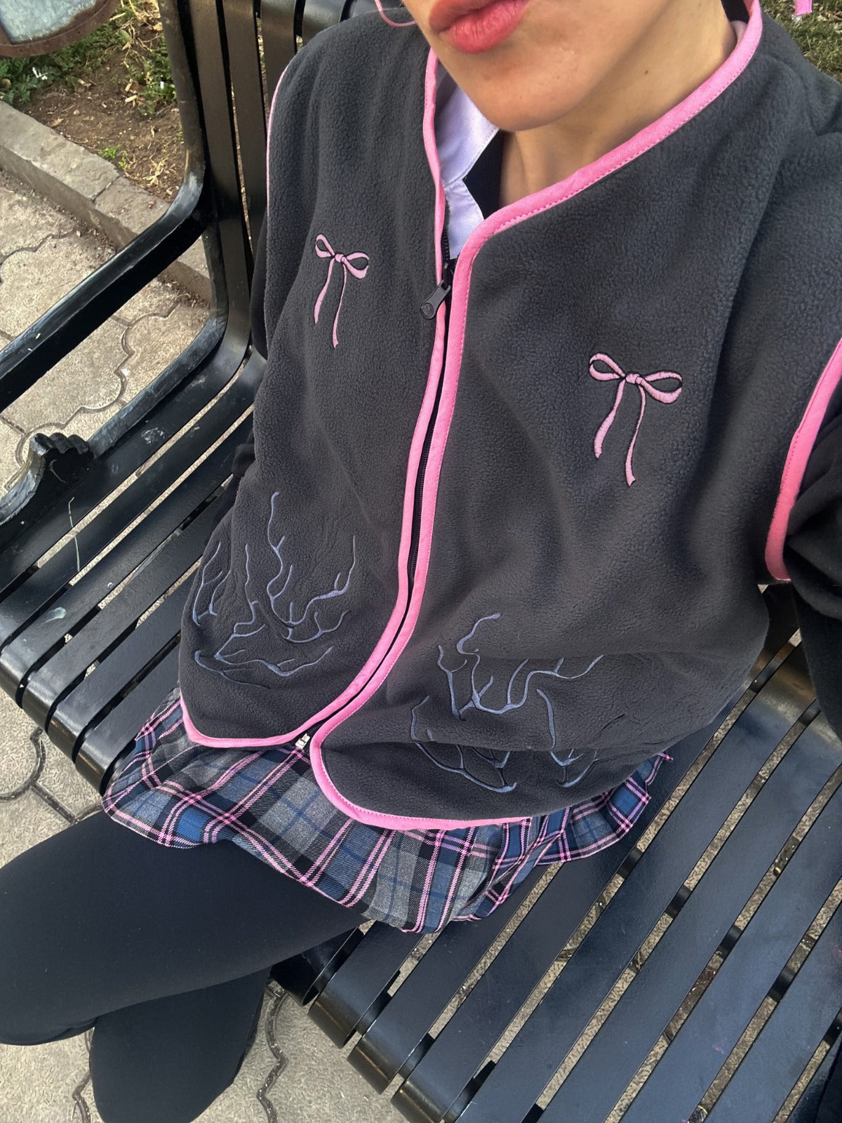 Person in gray jacket with pink bows, plaid skirt, and black leggings sitting on a bench.