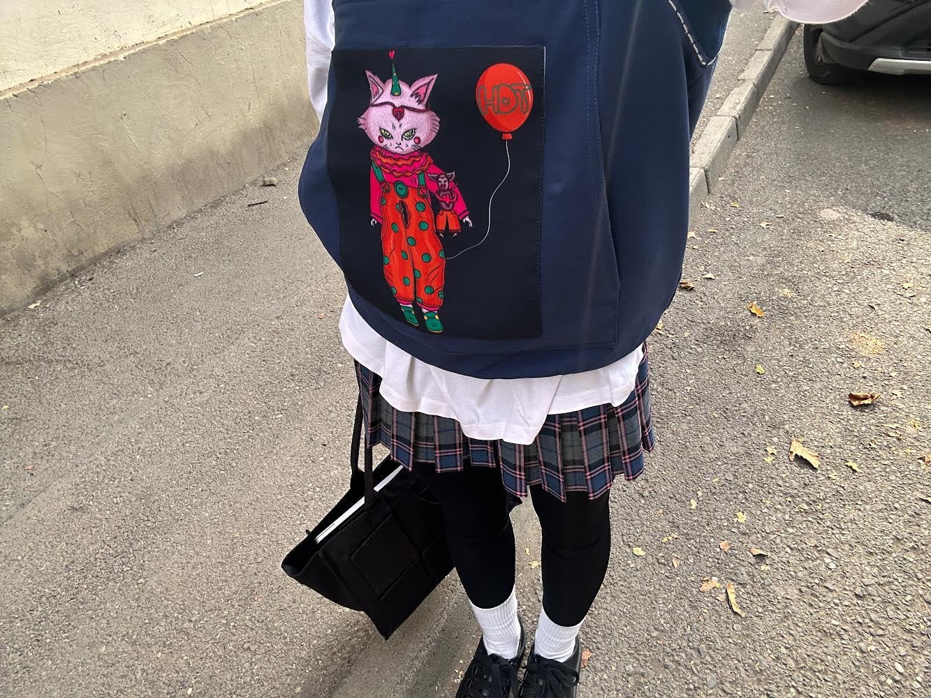 A person dressed in a plaid skirt, white shirt, and layered black leggings holds a black tote bag, wearing a navy vest featuring a playful cat graphic in a red outfit holding a balloon, adding a quirky and bold streetwear touch.