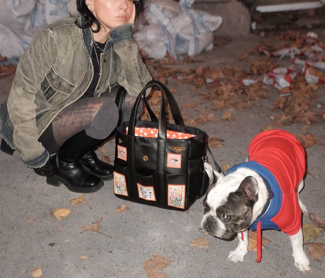 A black faux leather tote bag with colorful patchwork pockets sits on the ground next to a French Bulldog wearing a red hoodie, while a person crouches nearby in a green denim jacket and black boots.