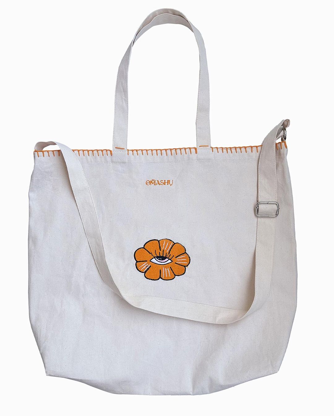 A white canvas tote bag with stitching, featuring a flower with eye inside. The bag has two sets of straps, one for over the shoulder and one for hand carrying. text above OMASHU