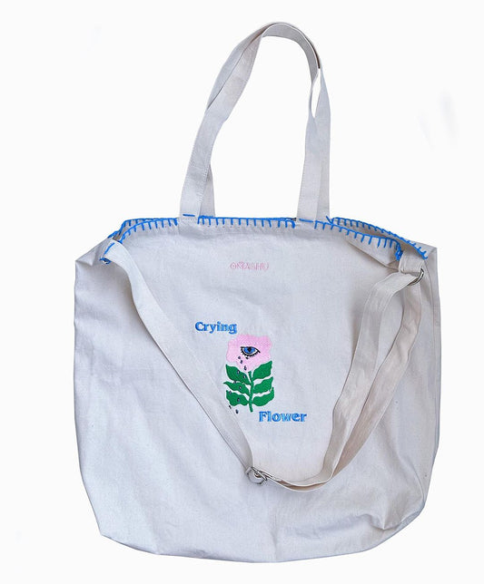 A white canvas tote bag with blue stitching, featuring a small pink and green graphic of a flower with the words ‘Crying Flower’ above it. The bag has two sets of straps, one for over the shoulder and one for hand carrying.
