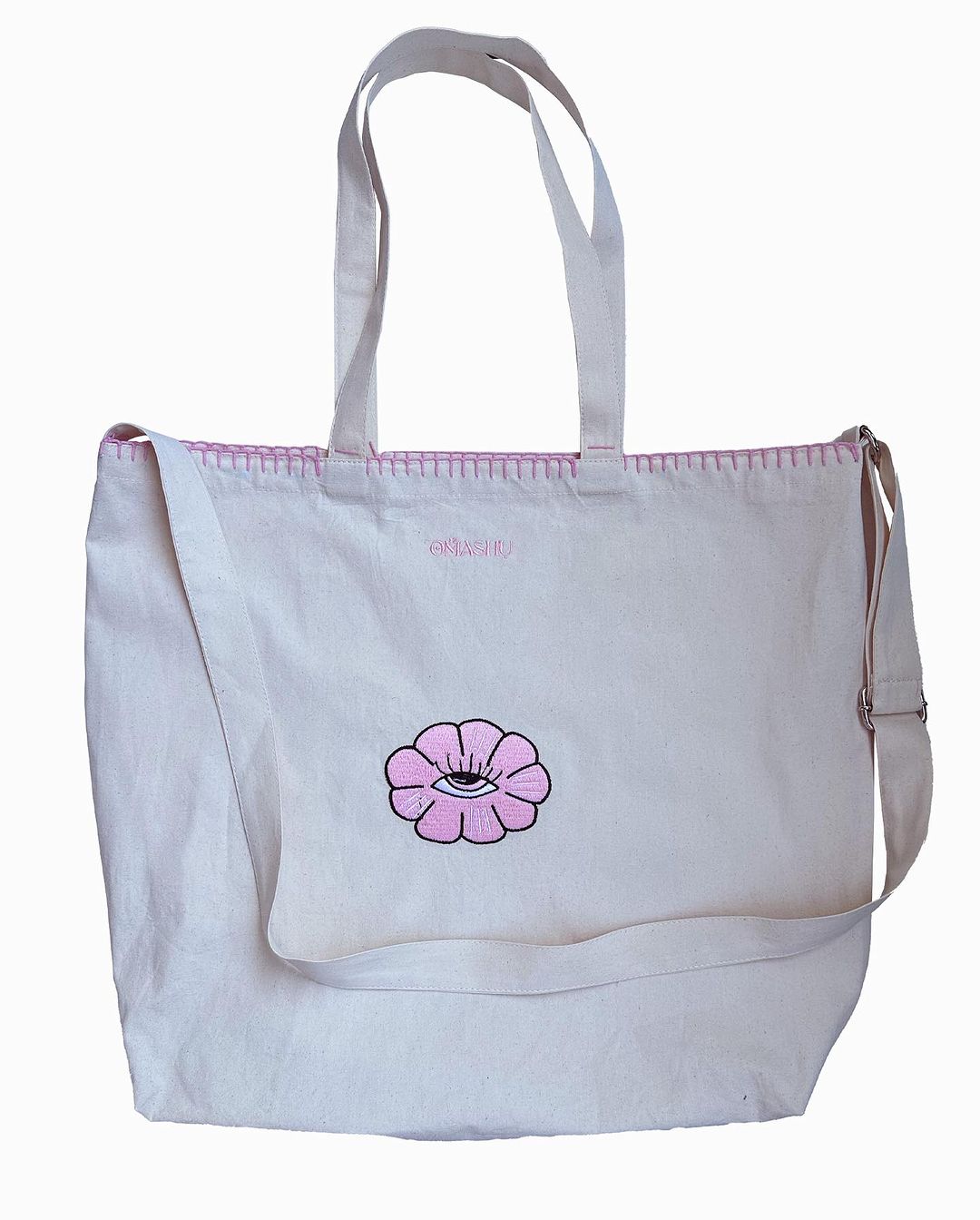 A white canvas tote bag with stitching, featuring a flower with eye inside. The bag has two sets of straps, one for over the shoulder and one for hand carrying. text above OMASHU