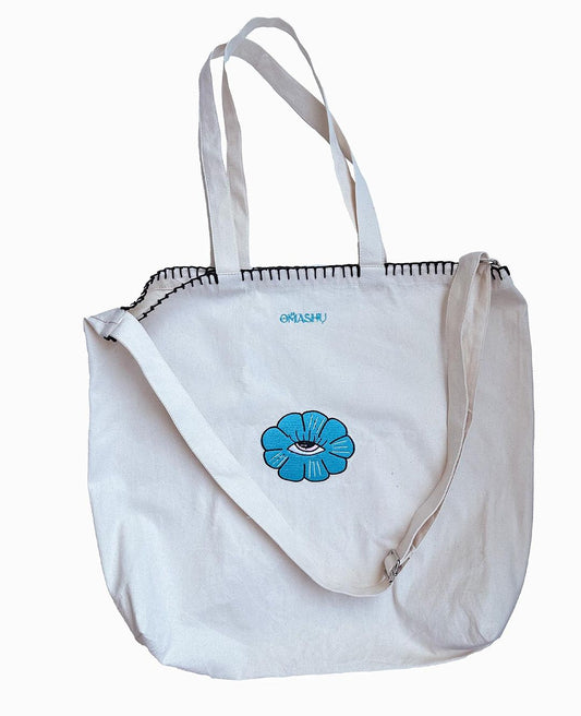 A white canvas tote bag with stitching, featuring a flower with eye inside. The bag has two sets of straps, one for over the shoulder and one for hand carrying.