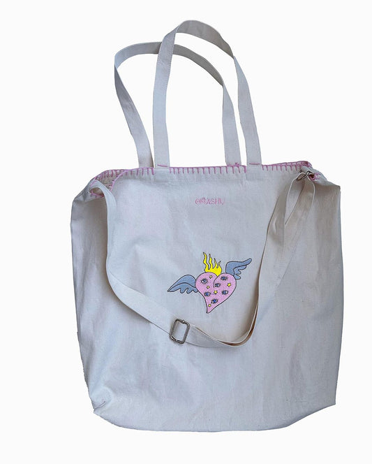  tote bag with a pink and blue design on it. The design is of a heart with wings and a crown on top. The heart is pink and the wings and crown are blue. The bag has two white straps and a silver buckle on the front