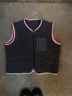 Gray vest with pink trimmings and zippered pockets laid on a concrete surface, accompanied by a pair of shoes.