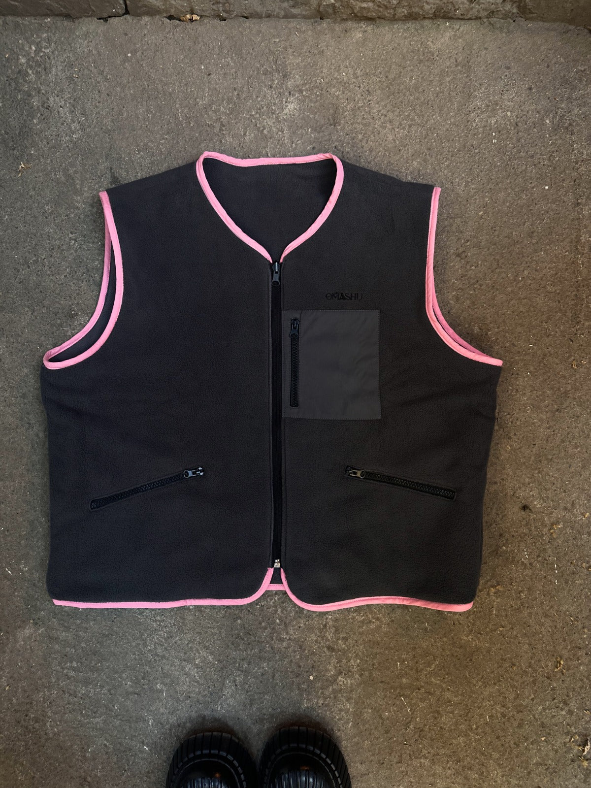 Gray vest with pink trimmings and zippered pockets laid on a concrete surface, accompanied by a pair of shoes.