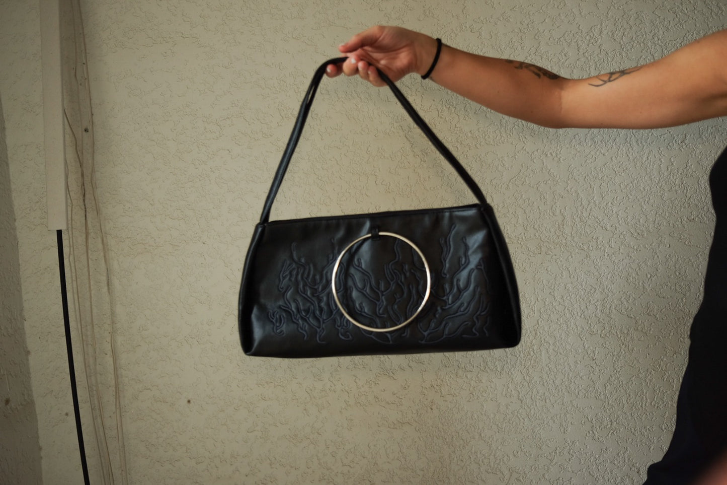 black handbag features a circular metallic handle. The bag’s surface showcases an embossed pattern, adding to its sophistication. Against the neutral-colored wall.