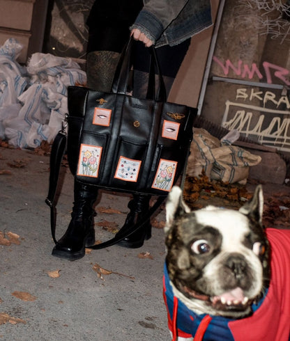 The black faux leather patchwork tote bag being carried by its handles, with colorful details visible, set against an urban backdrop of fallen leaves and graffiti.