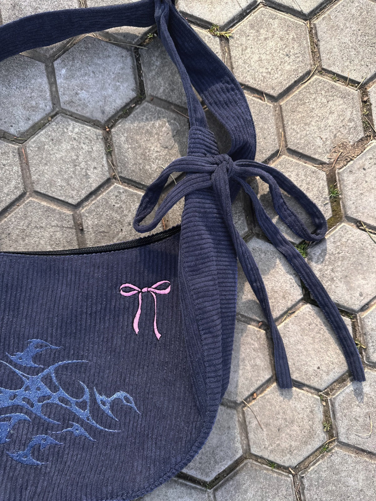 A close-up of the side of a gray corduroy bag, showing one of the tied knots securing the wide strap and a pink embroidered bow on the bag’s surface. The fabric’s texture and the intricate knot and embroidery details are highlighted.