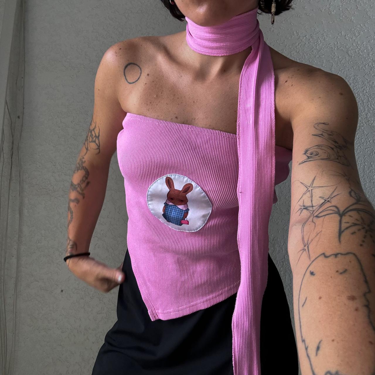 A person wearing a pink top featuring an embroidered patch of a cartoon-style bunny in a blue checkered outfit. The person is also wearing a matching pink scarf wrapped around their neck. Their arms are visible, showing several tattoos. The image is a close-up shot, focusing on the bunny patch and the upper body of the person.