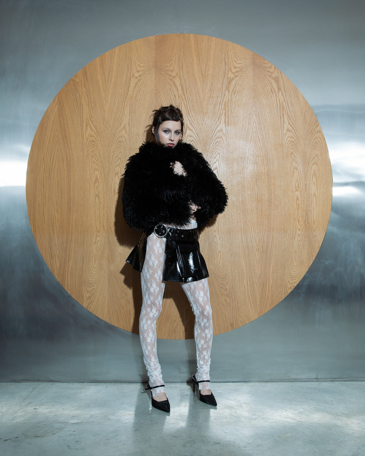 Model wearing the Open Front Charm Belt Skirt layered over lace tights, paired with a statement black furry jacket, standing confidently against a wood and metal backdrop.