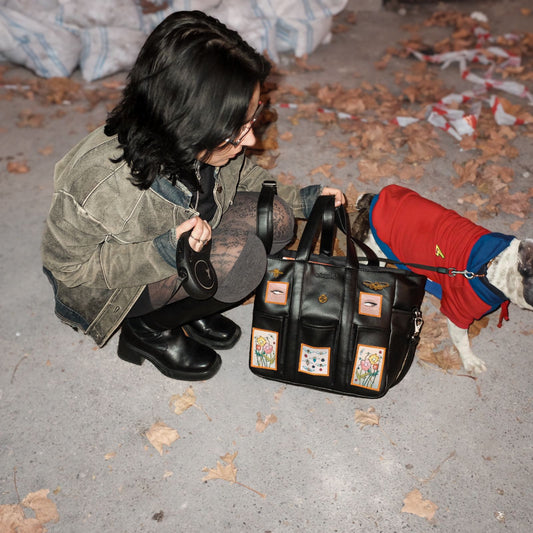 Close-up of the black patchwork tote bag featuring vibrant designs, held by a person in a green jacket and patterned tights, next to a French Bulldog in a red and blue outfit.