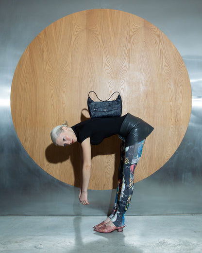 Blue noir embossed bag displayed with model bending forward against circular wooden backdrop, wearing black outfit