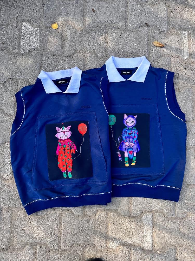Two navy sleeveless vests laid side by side, each with a whimsical cat graphic patch. One cat wears a red clown outfit holding a red balloon, and the other a blue hoodie with a green balloon, emphasizing playful and graphic streetwear.