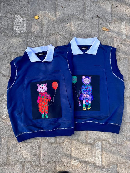 Two navy sleeveless vests laid side by side, each with a whimsical cat graphic patch. One cat wears a red clown outfit holding a red balloon, and the other a blue hoodie with a green balloon, emphasizing playful and graphic streetwear.