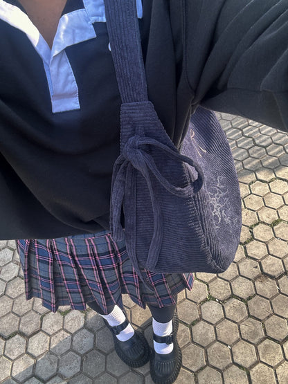 A person wearing a black shirt with omashu bag on shoulkder with bow, paired with a plaid skirt, standing outdoors.