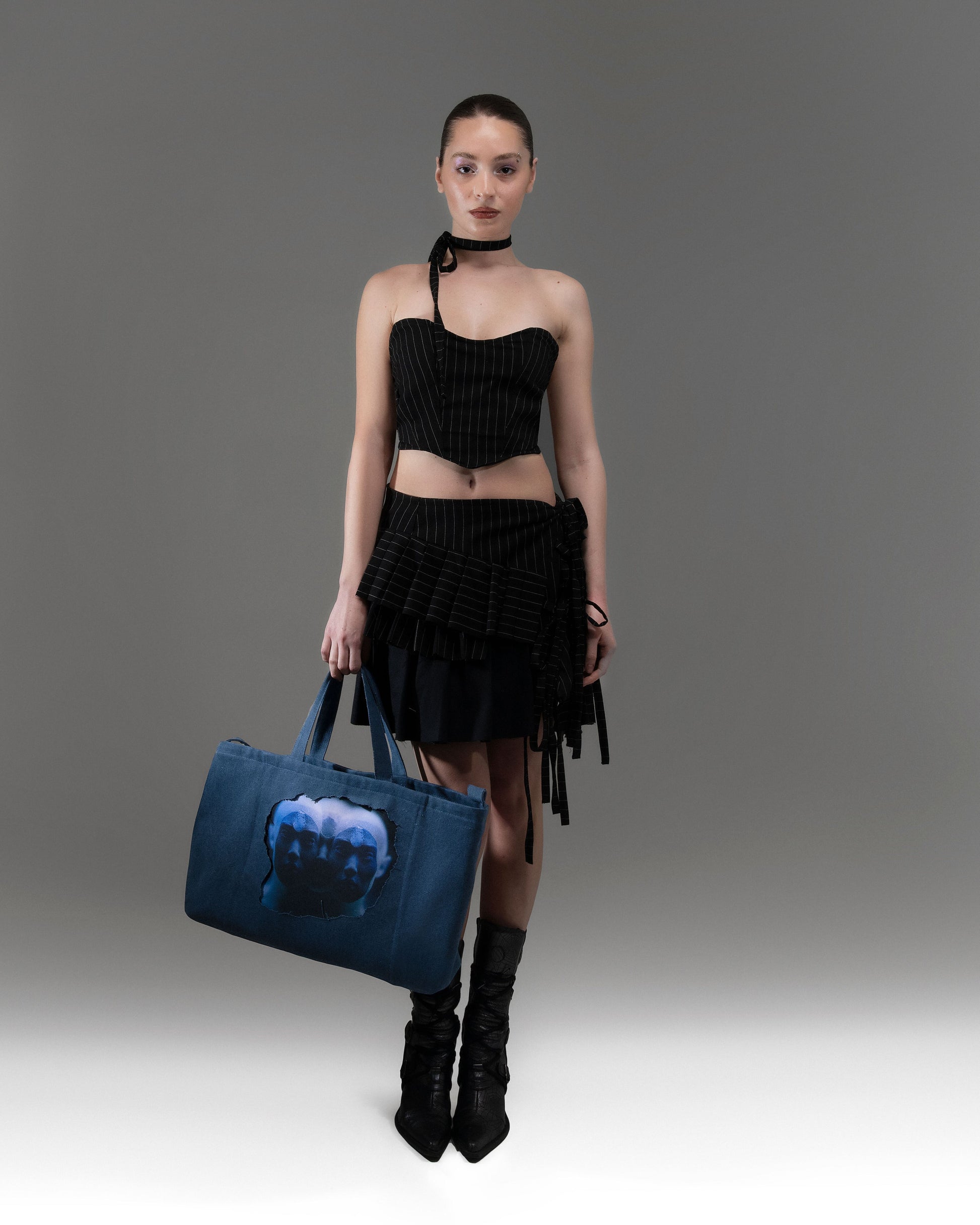 A person wearing a black outfit and holding a blue bag with an illustration, stands against a grey background.