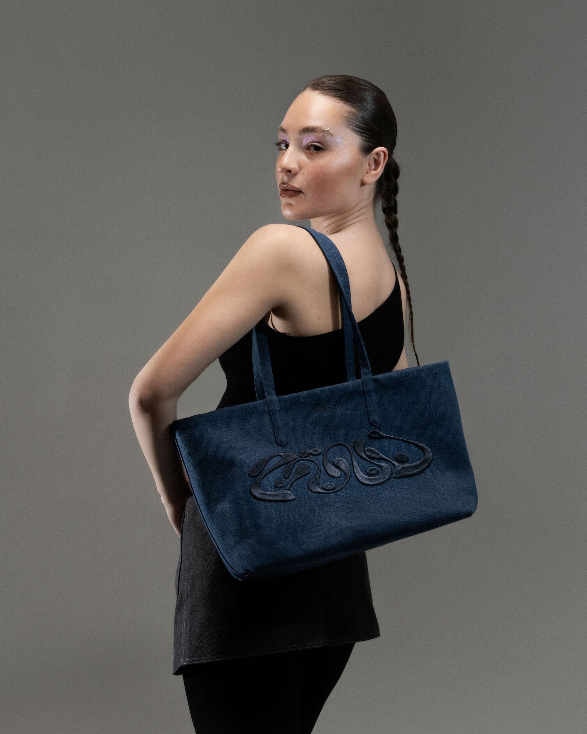 A person from a back and side angle, wearing a black sleeveless top and a braid, holding a large blue tote bag with an intricate design on the shoulder, against a plain grey background.