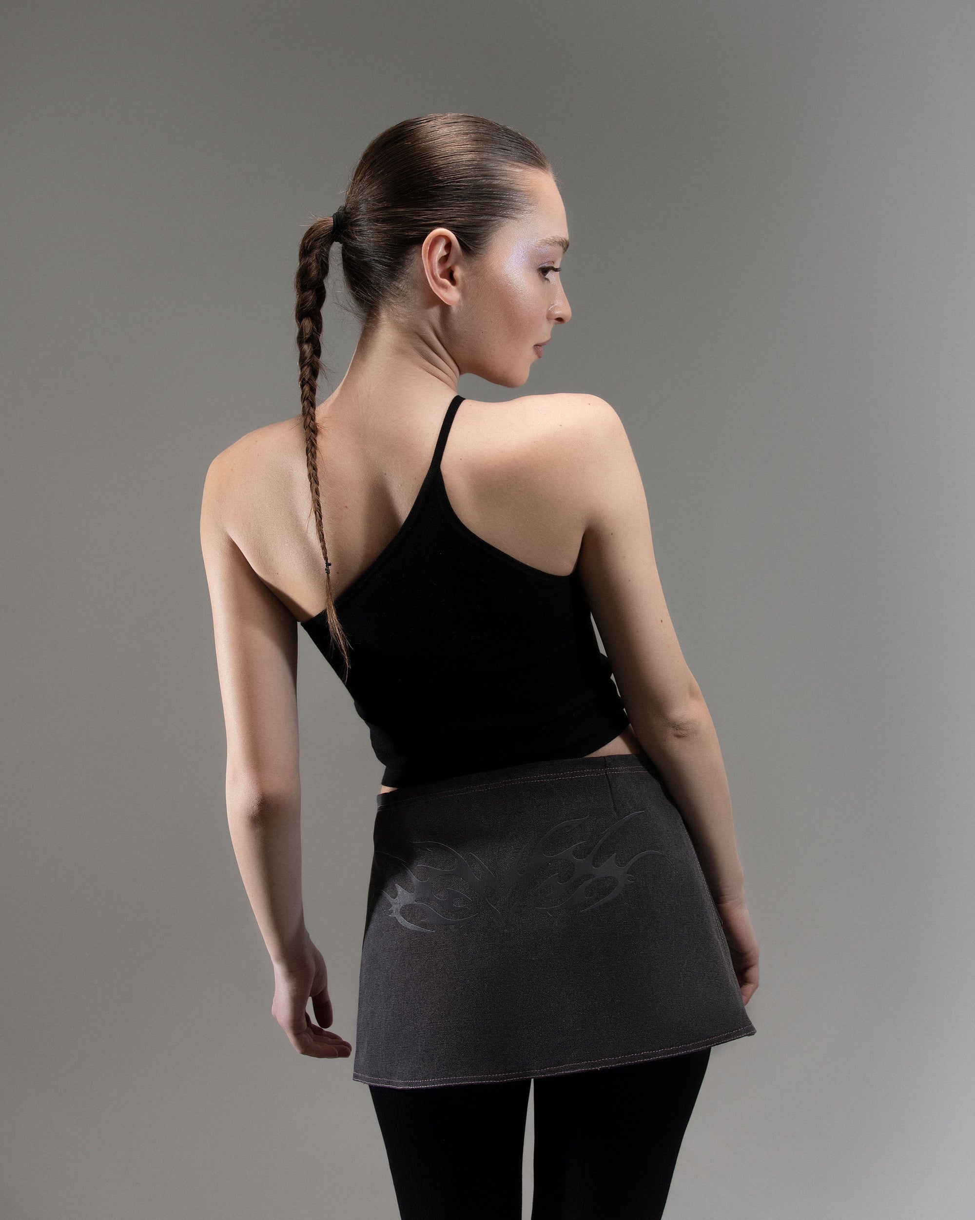 A person in a black sleeveless top and textured skirt stands against a grey background and hair tied in a neat braid down the back.