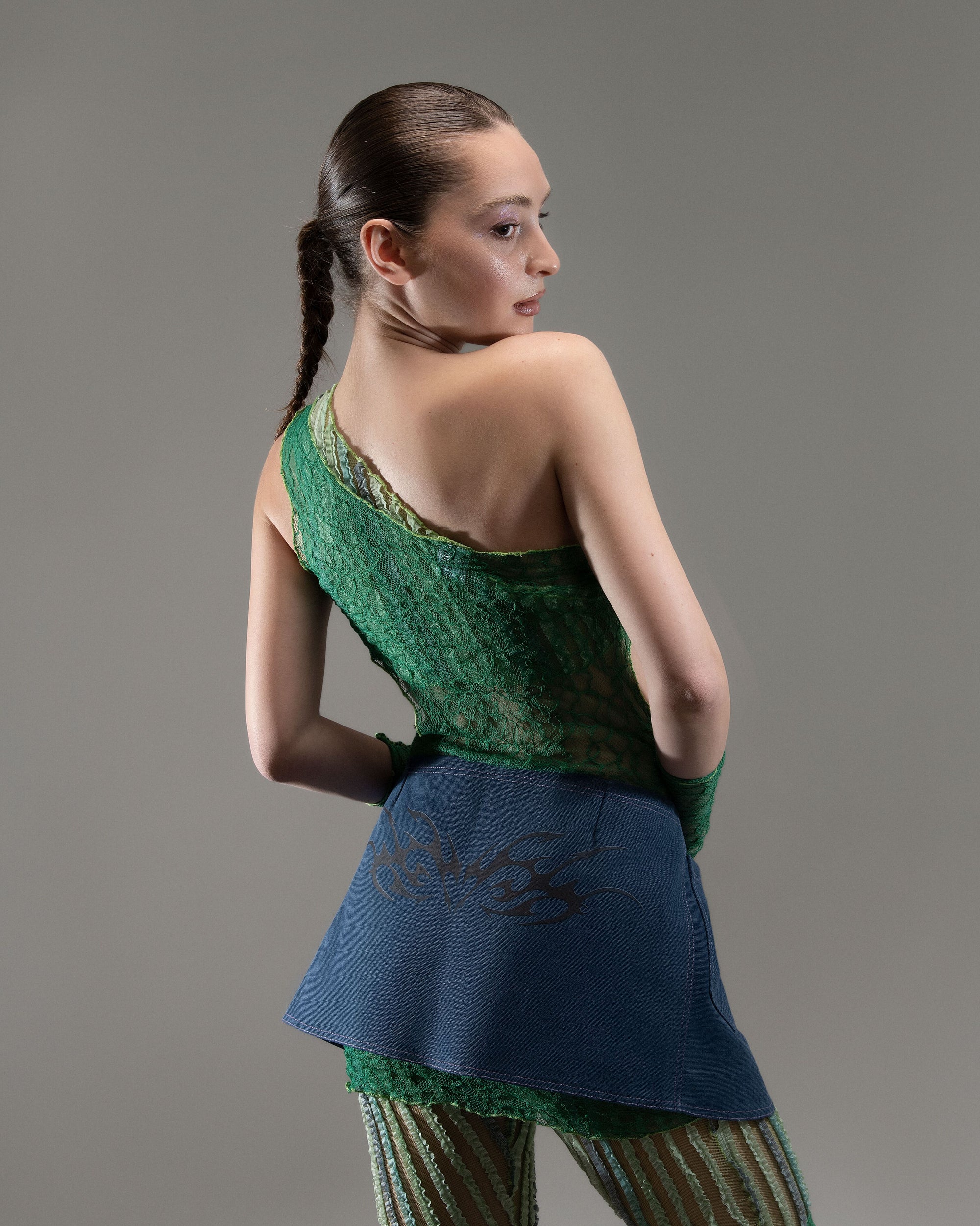 A person in a green sleeveless top and blue skirt poses against a grey background