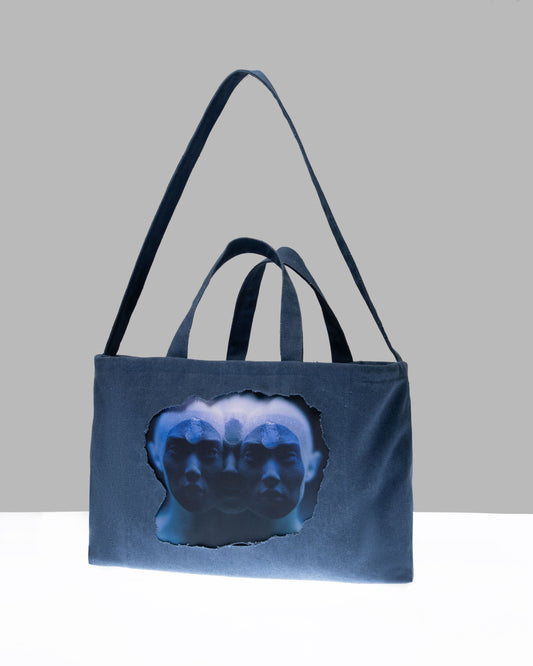 A blue tote bag with a cut-out window revealing a printed illustration, partially obscurred, against a light grey background.