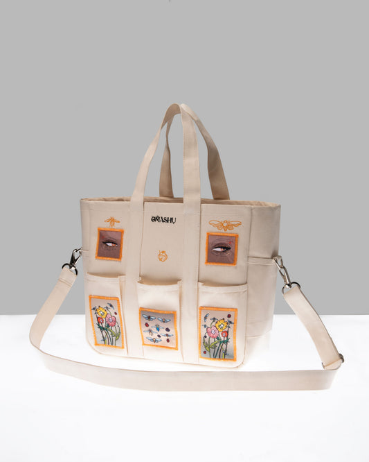A beige tote bag with multiple pockets, each adorned with different embroidered designs including flowers and a crown, and the word ‘OWASHI’ stitched at the top, against a grey background.