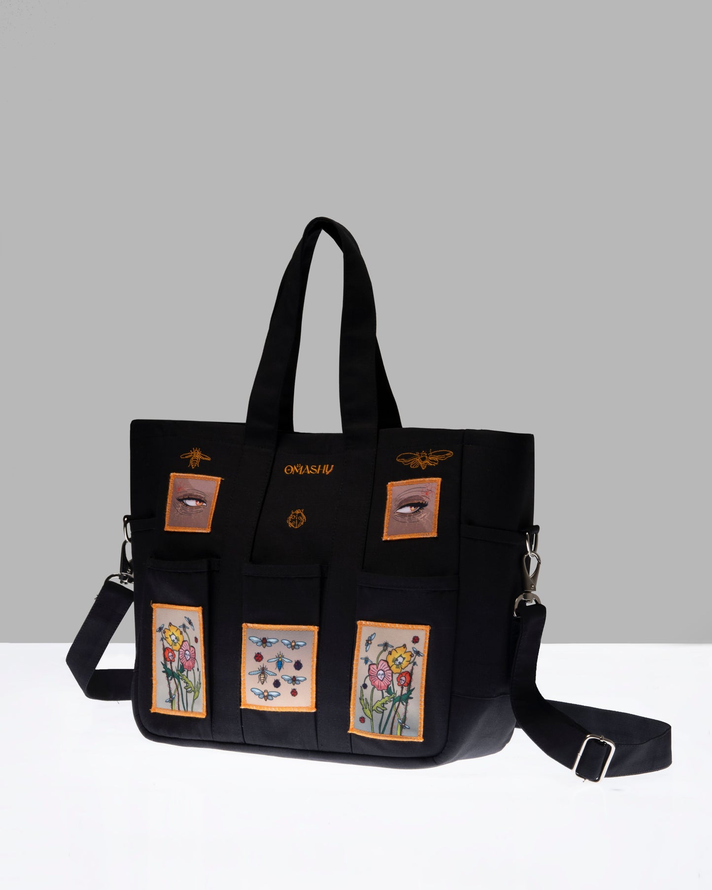 A black tote bag with embroidered floral designs and the brand name ‘OMASHU’ stitched on it, displayed against a grey background.