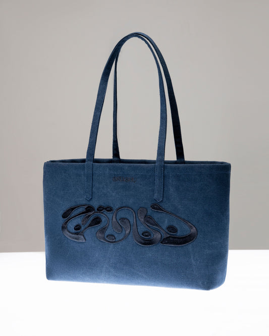 A stylish blue denim tote bag with dark handles, featuring an elegant black design on the front, placed upright on a white surface against a neutral background.