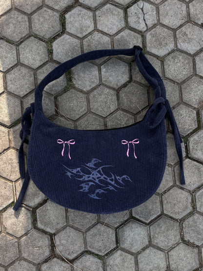 A stylish gray bag with intricate light blue embroidery and pink bow details, laid on a hexagonal paved surface