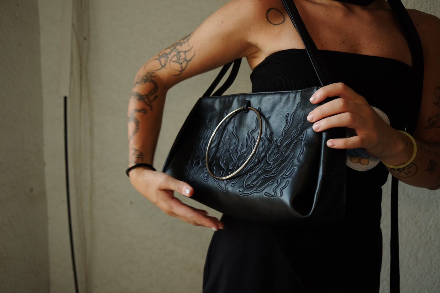 black shoulder bag features a circular metallic handle. The bag’s surface showcases an embossed or textured design, adding to its allure. Against the plain background.