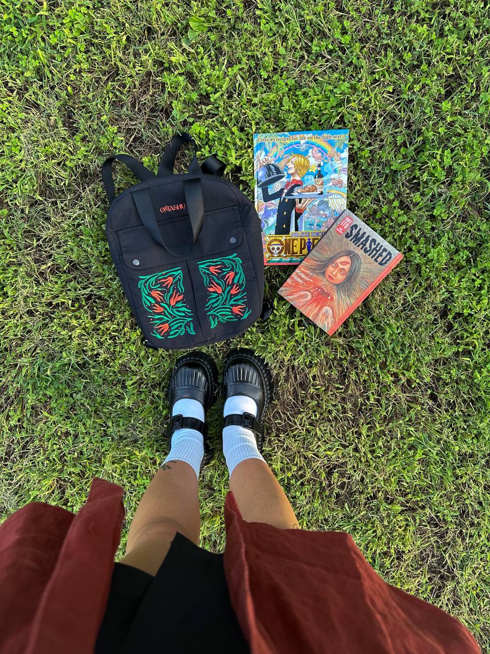 A person’s feet in black sandals and white socks and a black OMASHU backpack with a green and red floral pattern on a grassy ground with two comic books