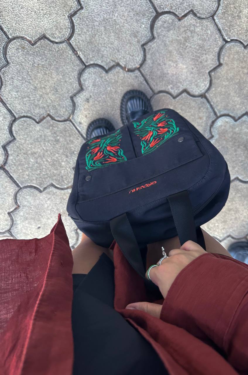 A person’s feet in black sandals with white stripes and a black OMASHU backpack with a red and green floral design on the front pocket on a gray pavement with a geometric pattern