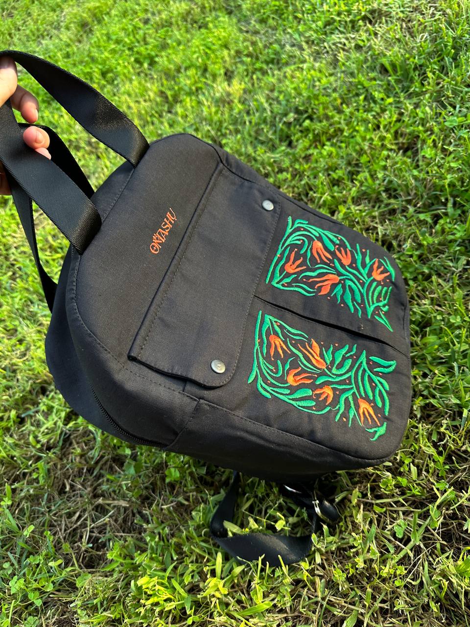 A black Omashu backpack with a black handle and straps and a colorful orange and green floral design on the front pocket being held up by a hand with grass in the background