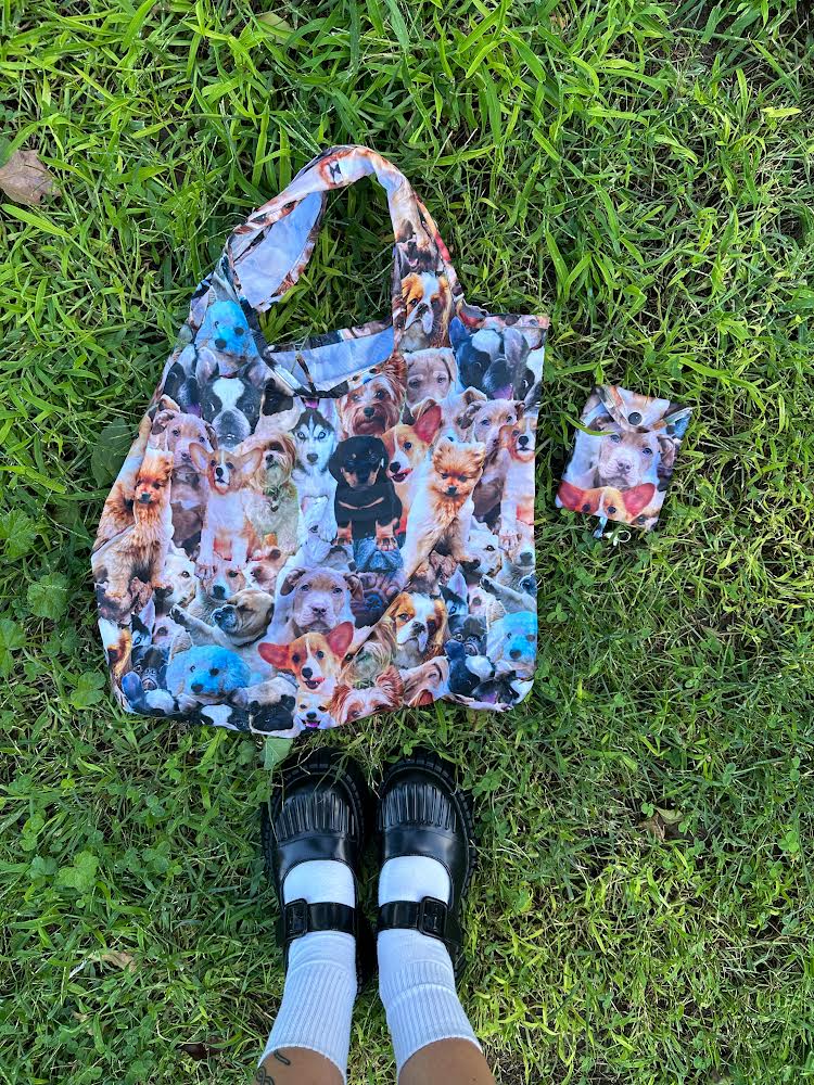 A tote bag with a colorful print of dogs, along with a small pouch, resting on a grassy lawn. The tote bag has two handles and is made of lightweight fabric, while the pouch has a zipper closure.