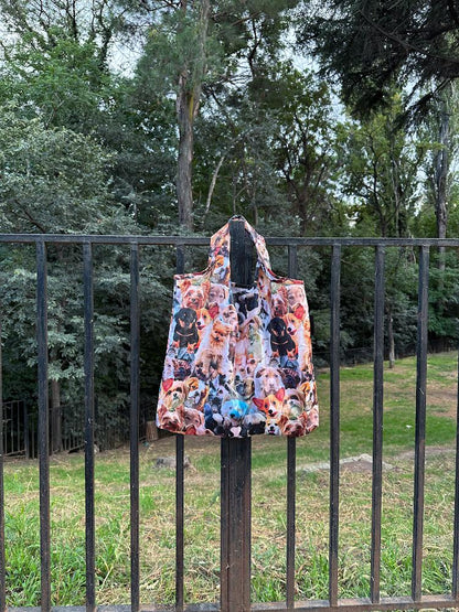 A tote bag with a colorful dogs print on it, hanging on a black metal fence against a backdrop of trees and shrubs.