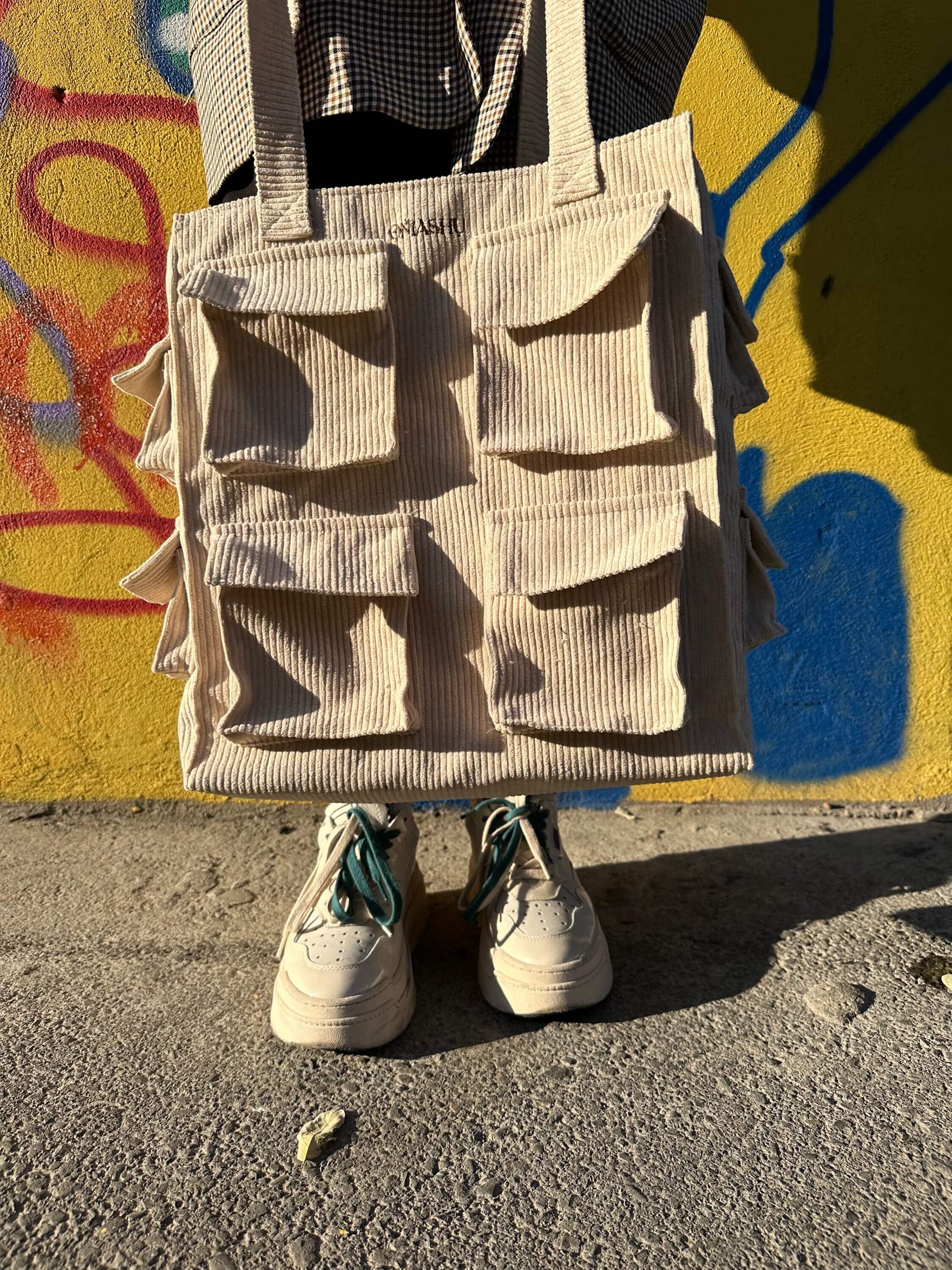 A beige corduroy tote bag with four pockets on the front and a pair of white sneakers with teal laces. The bag is hanging on a yellow wall with graffiti in the background. The sneakers are on the ground in front of the bag. The photo is taken in natural light and the shadows are visible.