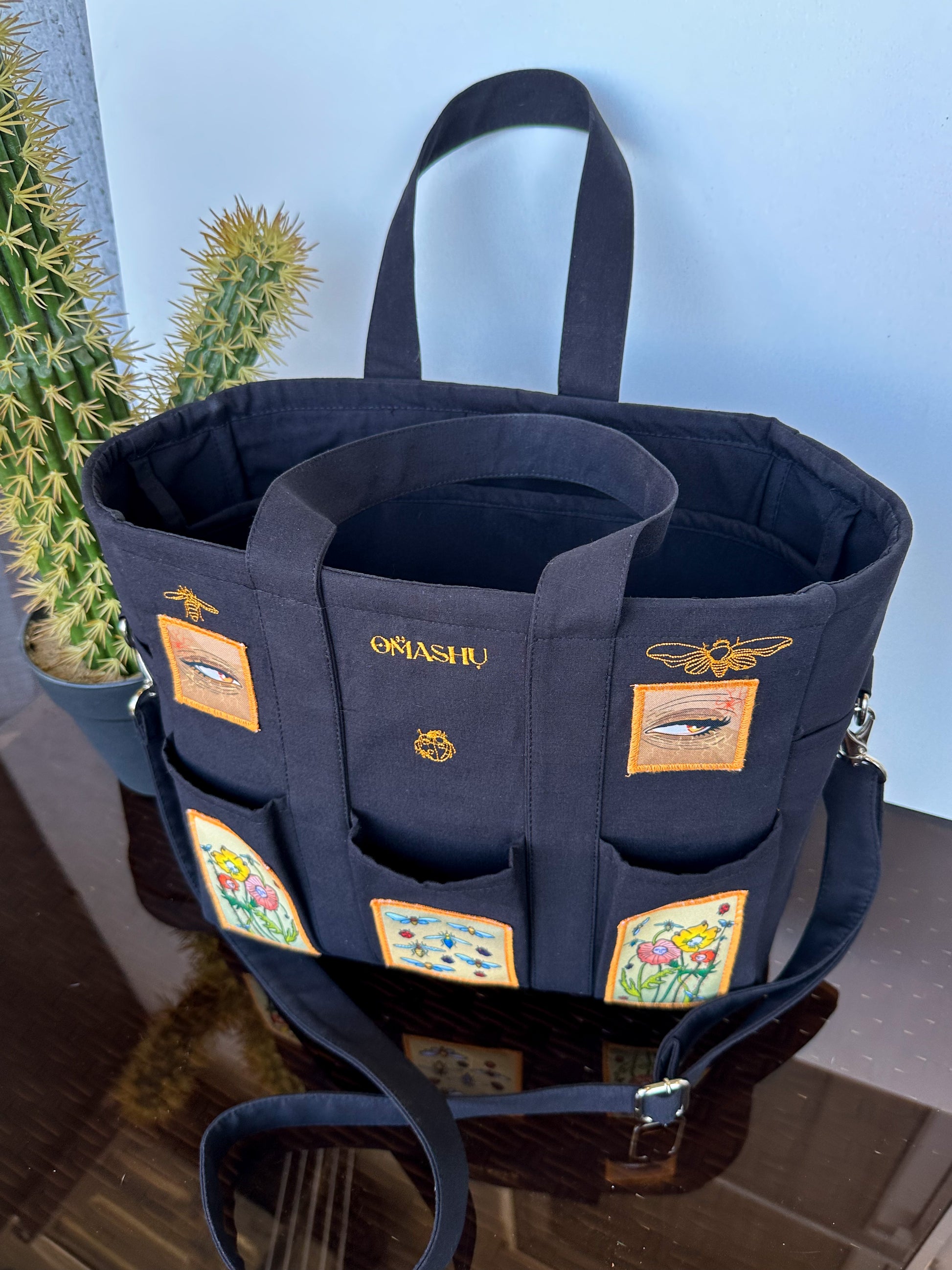 OMASHU black tote bag with multiple pockets on the front and sides with gold embroidered designs of bees, flowers, and a eye. The bag has two black handles and a black shoulder strap. The bag has a gold embroidered logo that reads ‘OMASHU’ on the front.