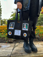 Black tote bag with various patches and pins arranged in a grid-like pattern. The patches include a cartoon polaroids and a receipt. The pins are round and have orange and yellow flowers on them. The person holding the bag is standing on a sidewalk with a flower bed in the background.
