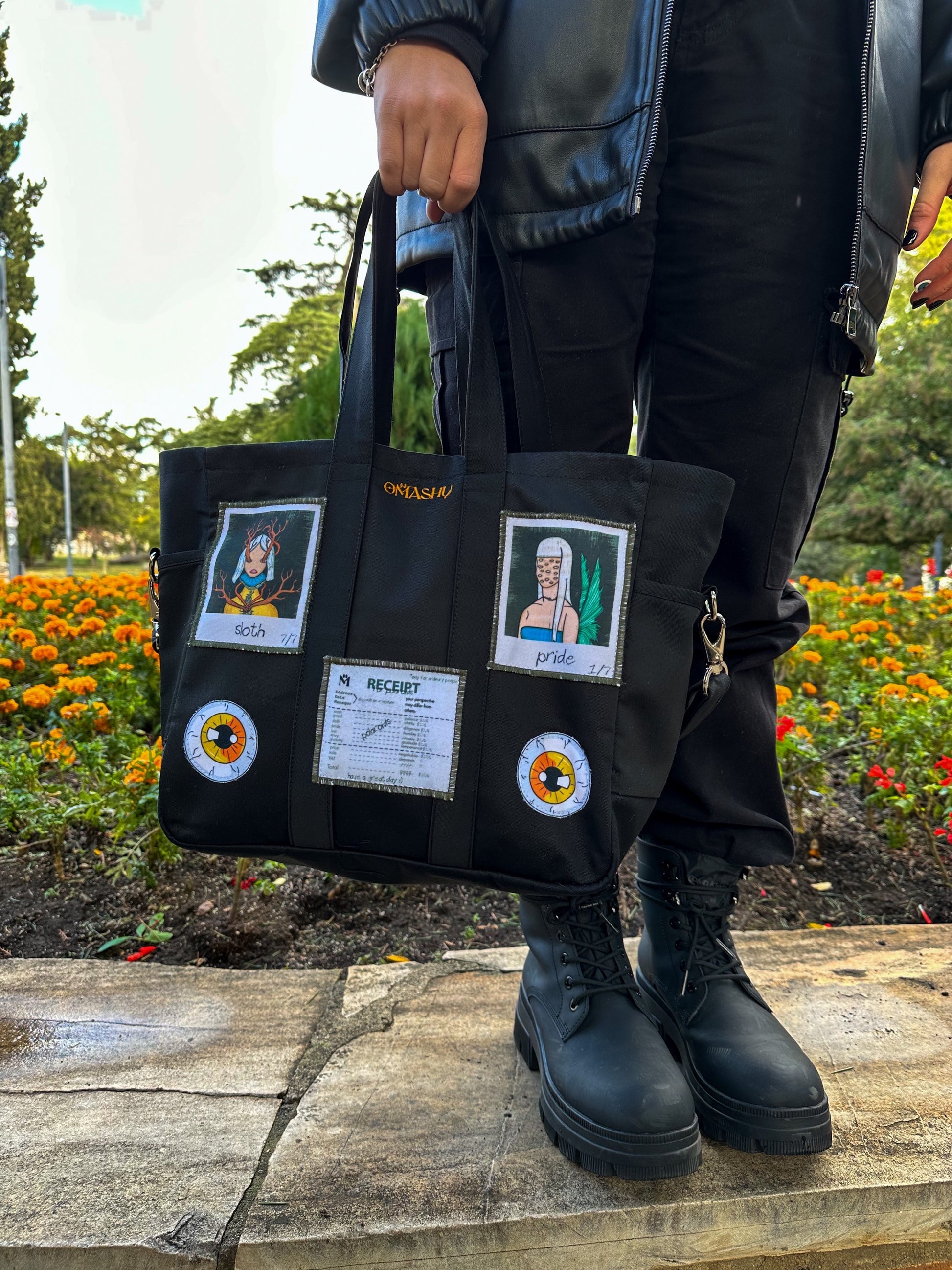 Black tote bag with various patches and pins arranged in a grid-like pattern. The patches include a cartoon polaroids and a receipt. The pins are round and have orange and yellow flowers on them. The person holding the bag is standing on a sidewalk with a flower bed in the background.