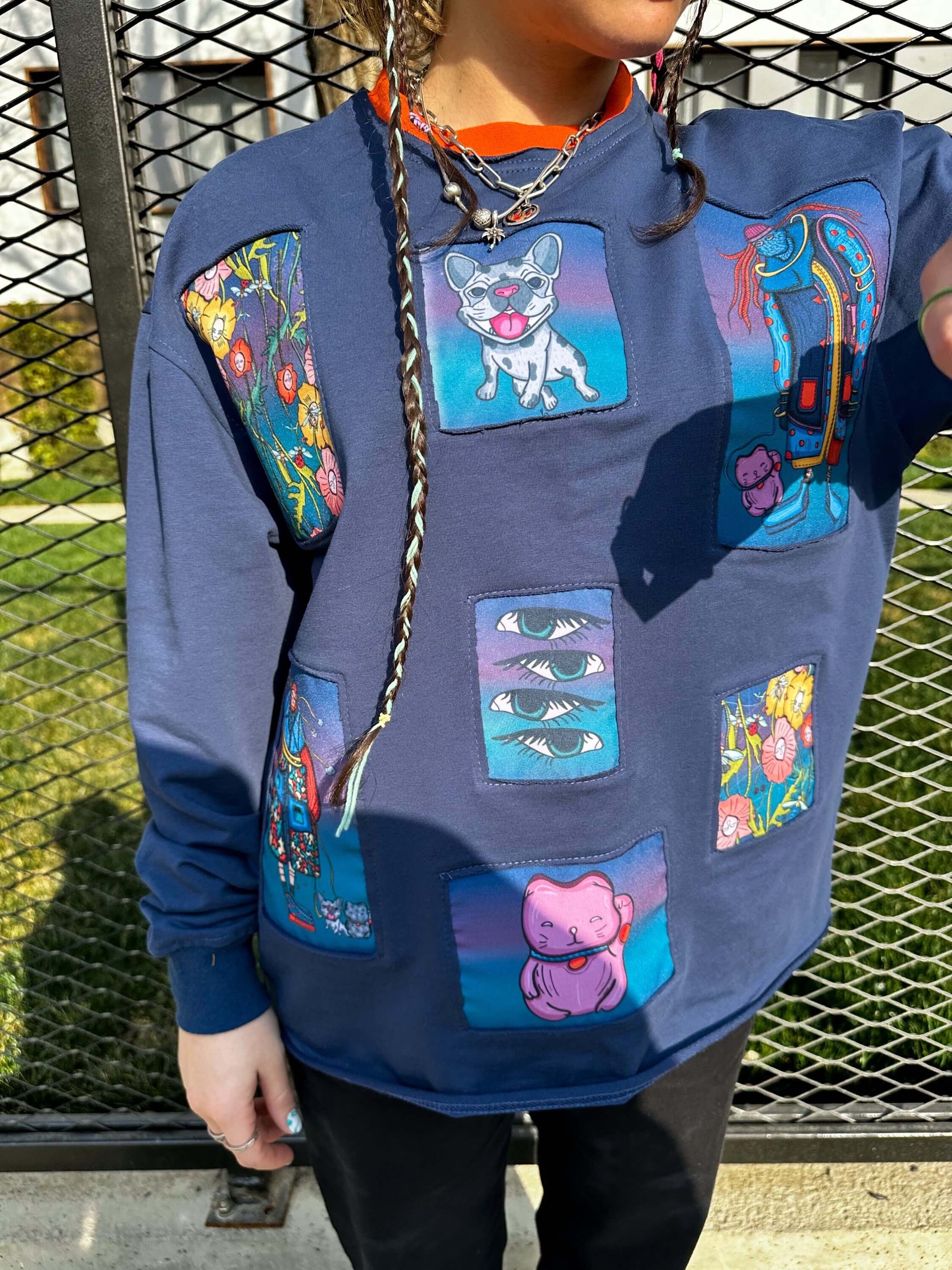 A photo of a person wearing a blue sweatshirt with a crew neck and long sleeves, featuring colorful cartoon-style illustrations of a cat, a dog, a bird, a flower, and a robot in various colors such as pink, blue, green, and yellow, with a necklace with a large pendant, against a chain link fence background with greenery behind it.