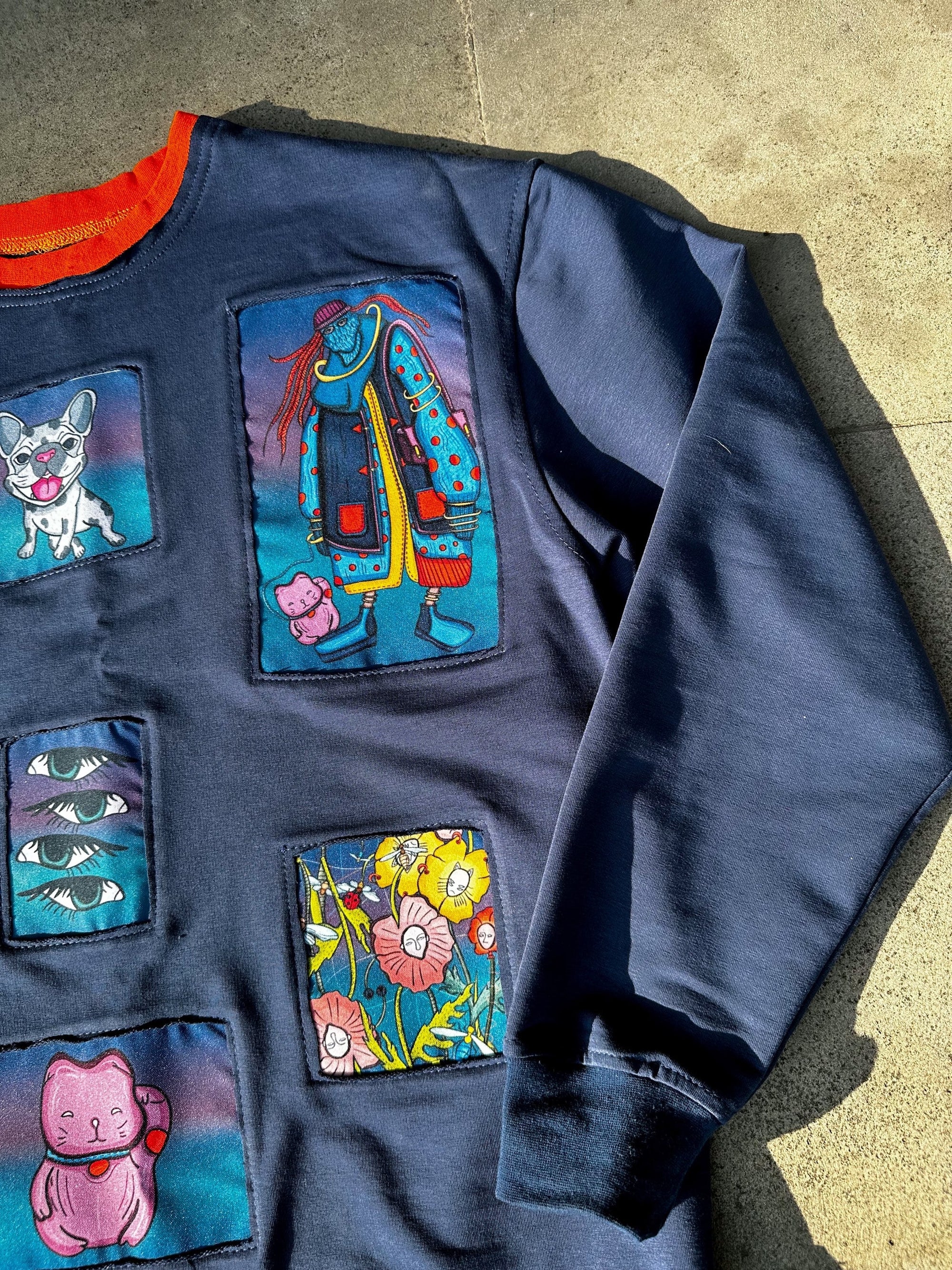 A navy blue sweatshirt with colorful patches in different sizes and shapes, depicting images such as a cat, a dog, a flower, and a person wearing a hat and coat, laid out on a concrete surface with sunlight shining on it, with orange collar.