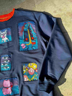 A navy blue sweatshirt with multiple colorful graphic patches, laid flat on a concrete surface. The patches feature whimsical illustrations including a dog, a character in a coat, eyes, flowers, and a pink cat.